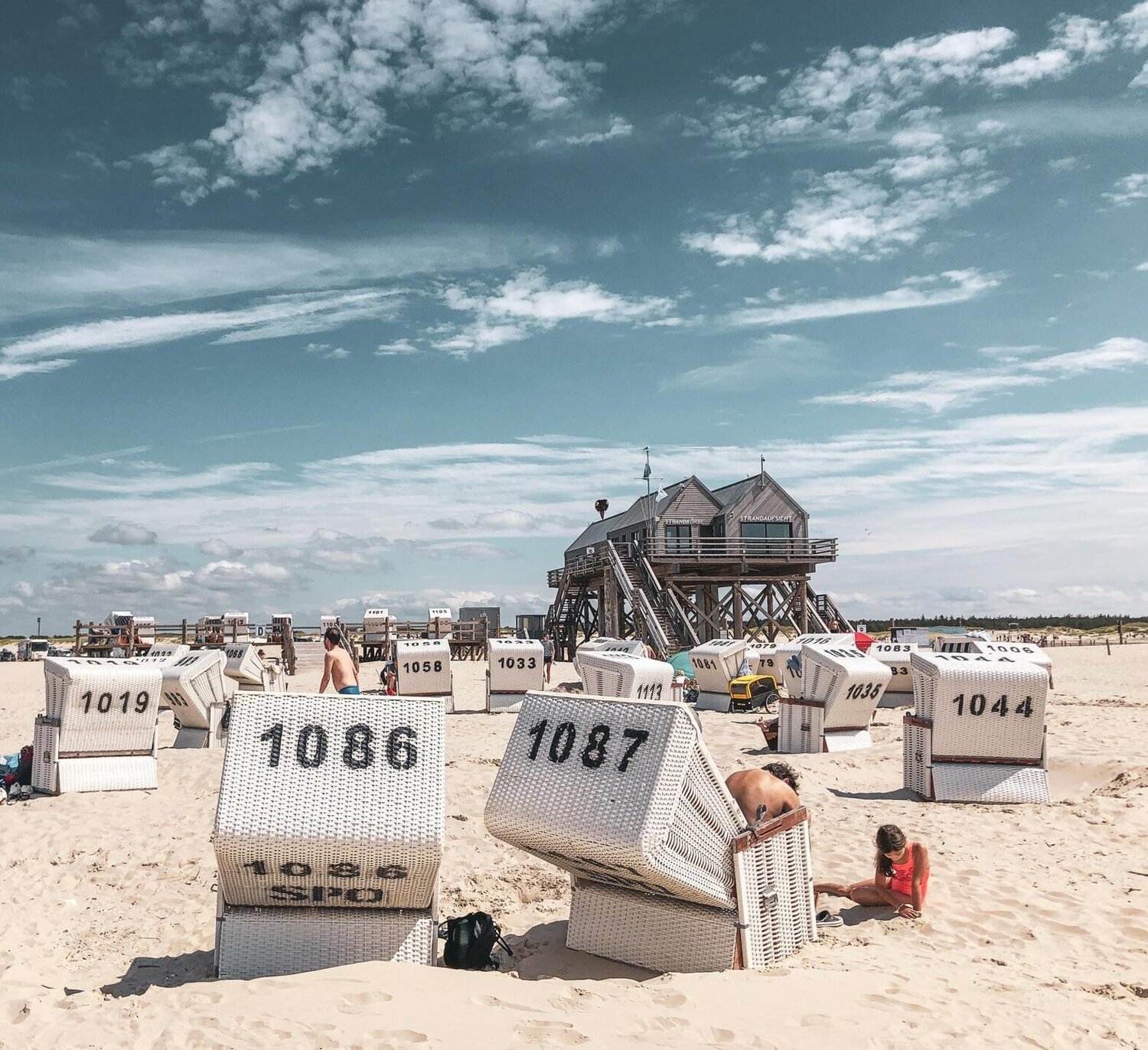 In den Strandkörben genießen die Strandbesucher die Sonne. Im Hintergrund steht ein Pfahlbau.