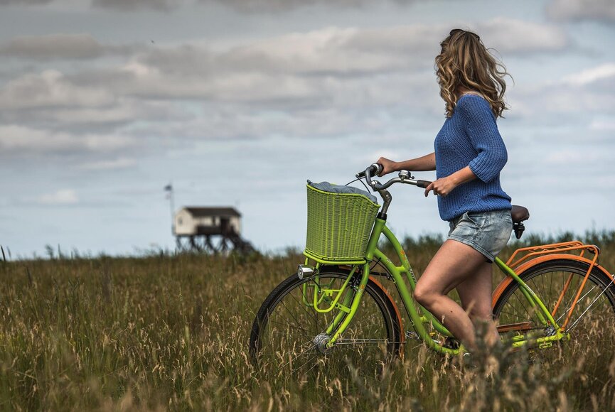 Frau mit Fahrrad steht in den Salzwiesen, Pfahlbau im Hintergrund
