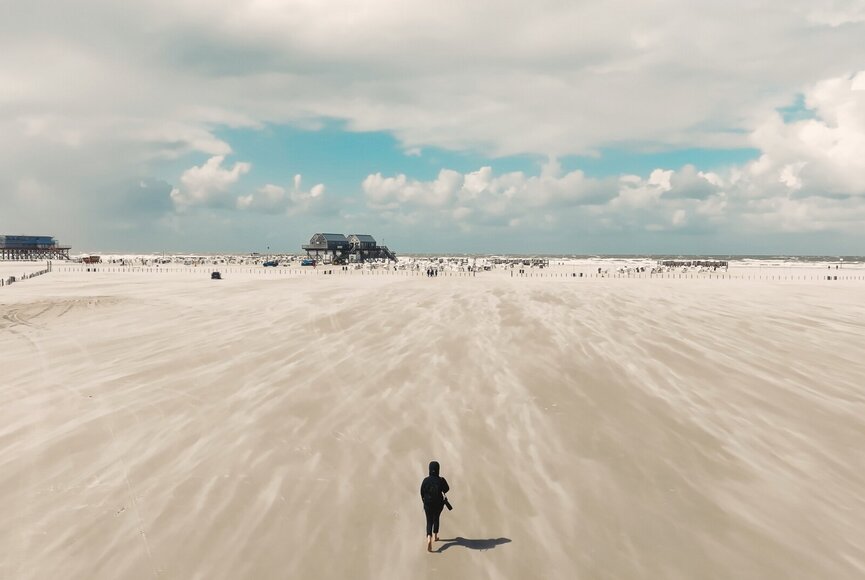 Strandbild mit viel Weite, man sieht den Sand im Wand fliegen, eine Person mit Kamera läuft am unteren Bildschirmrand ins Bild rein Richtung Meer